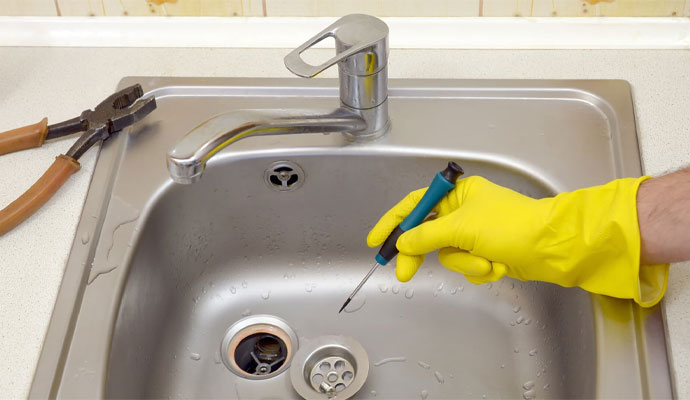 A hand wearing a yellow glove using a screwdriver to repair the drain strainer of a stainless steel kitchen sink