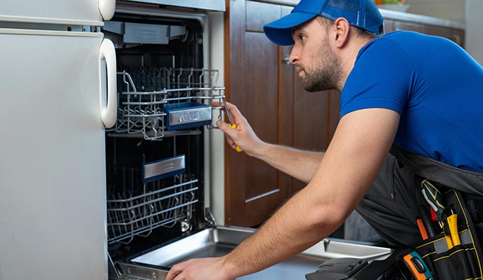 Technician in blue shirt and cap repairing an open dishwasher with tools on belt