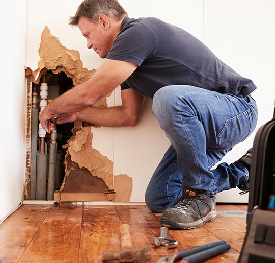 Person kneeling on a wooden floor repairing exposed pipes inside a damaged wall with tools nearby