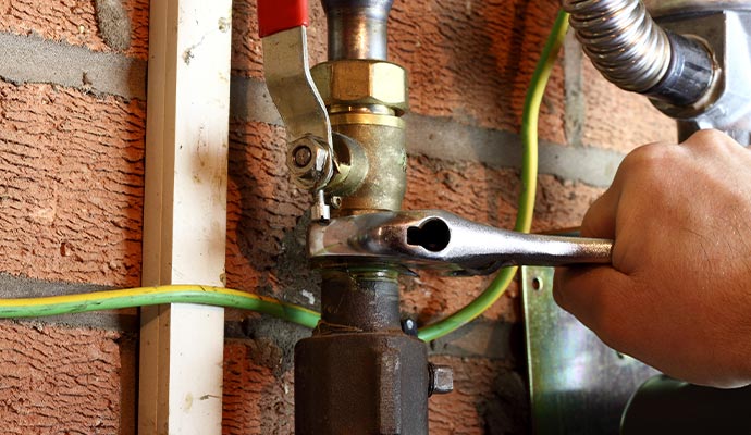 Technician using a wrench to adjust a valve on a gas pipe near a brick wall with visible wiring