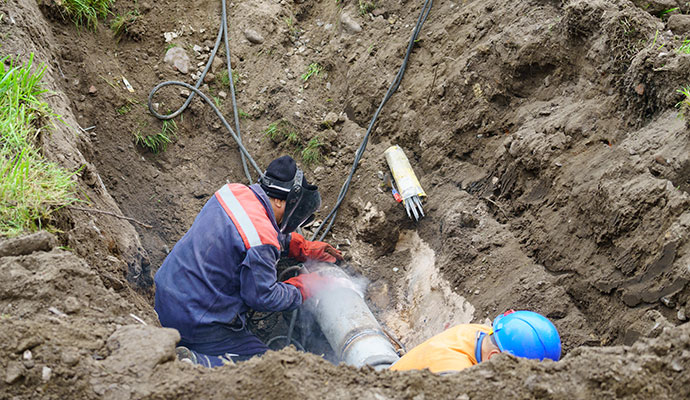 Two technicians working on a metal pipe inside a deep excavated trench